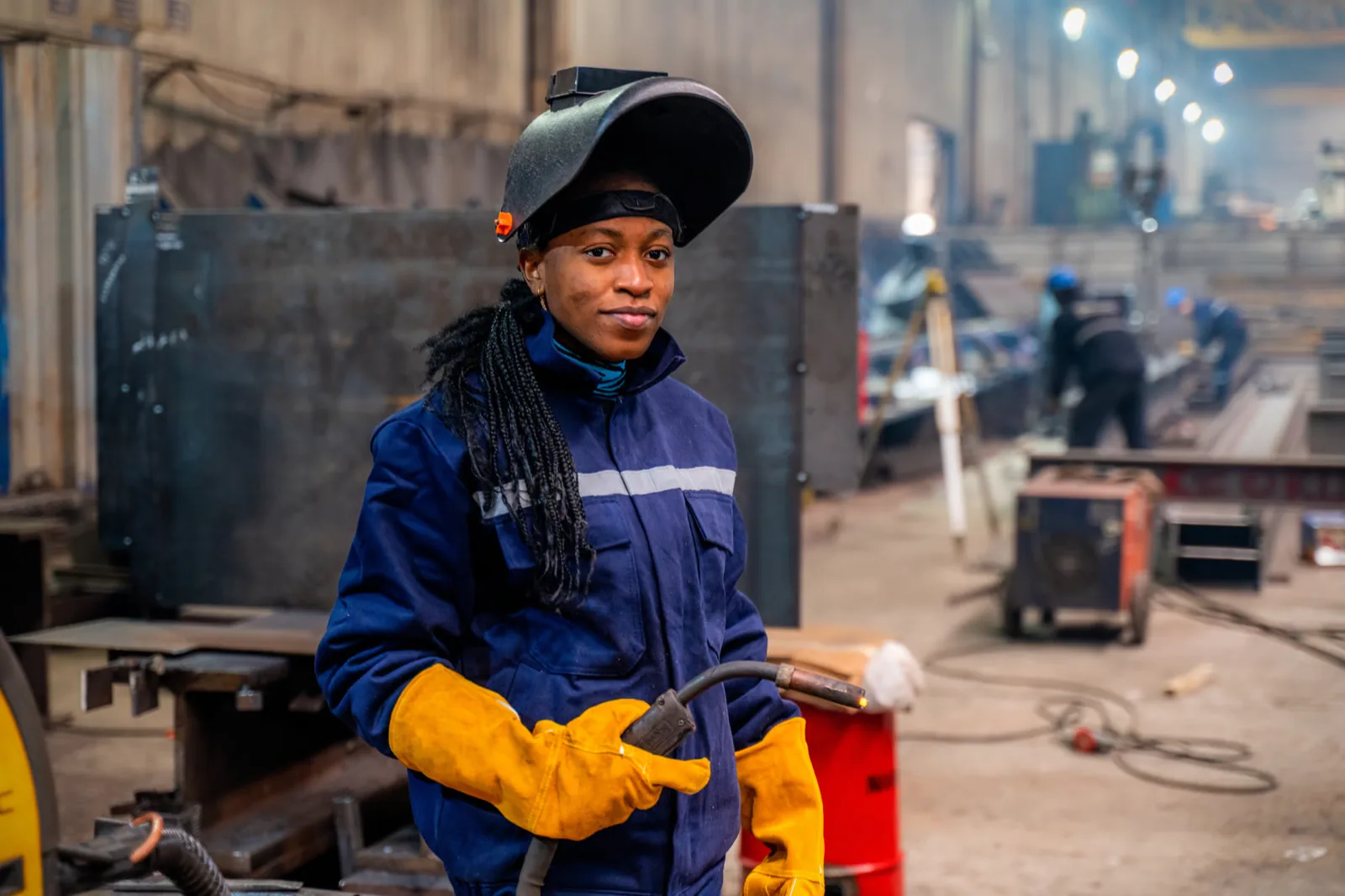 A young woman in welding coveralls holding a MIG torch, helmet flipped up